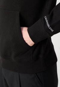 Sudadera negra con capucha, puños y bajo acanalados, con un bolsillo frontal. "CALVIN KLEIN JEANS" está bordado en blanco en el puño. Textura suave.