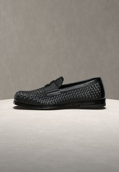 Mocassin en cuir noir avec texture tissée et détail de bride lisse sur une surface beige, devant un fond gris neutre.