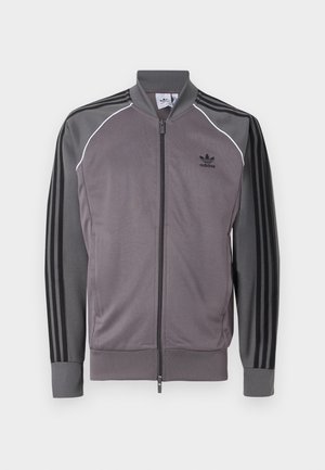 Szara sportowa kurtka z zamkiem z przodu, czarny wzór z trzema paskami na rękawach, ściągacze na mankietach i kołnierzu. Posiada białą lamówkę wzdłuż szwów.