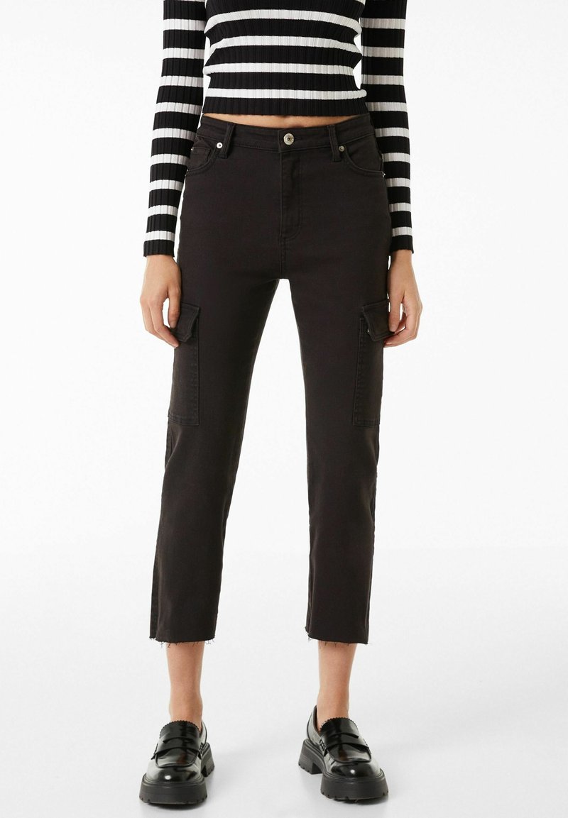 Pantalon cargo noir avec deux poches latérales avant, longueur raccourcie, ourlet effiloché et tissu robuste et texturé ; porté avec un haut rayé noir et blanc.