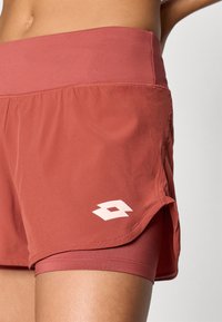 Lyhyet urheilushortsit punaruskeassa sävyssä. Valmistettu kevyestä kankaasta, jossa on joustava vyötärö ja valkoinen logotaito helman kohdalla.