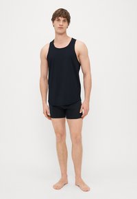 Črna majica na naramnice in črne kratke hlače iz mehkega materiala, z ohlapnim krojem in preprostim dizajnom. Model stoji bos na nevtralni podlagi.