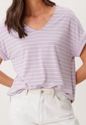 Mujer vistiendo una blusa de manga corta en mora claro con escote en V y rayas horizontales blancas, metida dentro de unos pantalones vaqueros blancos de cintura alta.