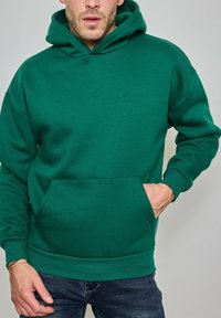 Homme portant un sweat à capuche vert uni avec poche avant, main dans la poche, et un jean bleu devant un fond neutre.