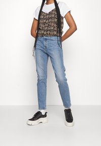 Svart mönstrad linne över en vit t-shirt, matchad med ljusblå jeans och svarta sneakers med en tjock vit sula. Flätat hår synligt.