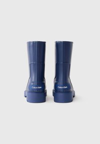 Bottes en caoutchouc bleu marine à chevilles avec une finition mate, arborant un design texturé et un logo blanc "Calvin Klein" à l'arrière. Bout carré.