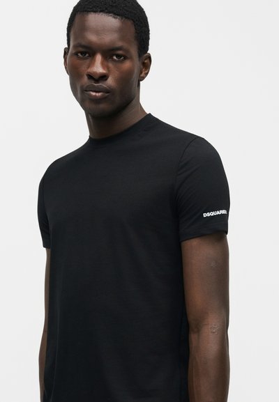 Άνδρας που φοράει απλό μαύρο κοντομάνικο μπλουζάκι με το κείμενο "DSQUARED" στο αριστερό μανίκι, στέκεται μπροστά από ένα απλό φωτεινό φόντο.