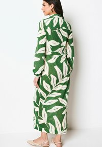 Robe longue verte avec motifs de feuilles blanches, en tissu léger, design à col, manches longues, associée à des sandales plates beiges.