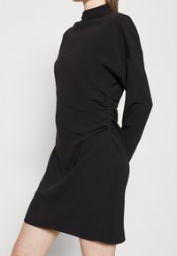 Robe noire à col montant, manches longues, avec fronces latérales, en tissu lisse et extensible. Longueur courte avec une silhouette ajustée.