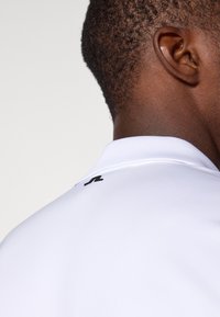 Hvit polo skjorte med en glatt tekstur, med en svart logo på kragen. Designet er minimalistisk, og fremhever enkelhet og komfort.