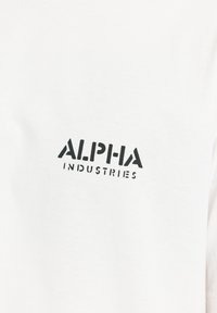 Λευκό ύφασμα με μαύρο κείμενο "ALPHA INDUSTRIES" τυπωμένο με έντονα κεφαλαία γράμματα, κεντραρισμένο στην περιοχή του στήθους.