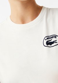 Närbild av en person som har på sig en vit t-shirt med en svart Lacoste-logotyp som föreställer en krokodil på vänster bröst.