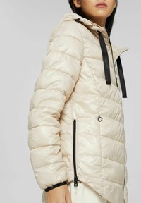 Chaqueta acolchada de color beige claro con acolchado horizontal, capucha, cordones de acento negros y bolsillos laterales con cremallera negra.