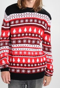 Osoba w czerwonej, białej i czarnej świątecznej bluzie z wzorami choinek i płatków śniegu, połączonej z szarymi dżinsami.
