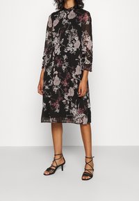 Woman wearing black midi dress with pink and white floral print, long sheer sleeves, and black strappy heeled sandals.