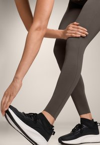 Szürke leggings sima textúrával, fekete sportcipőkkel, fehér talppal és texturált fekete felsőrésszel, dinamikus pózban.