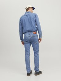 Camisa de denim azul de manga larga, con puños abotonados y una costura en el yugo trasero. Combinada con jeans azul claros y zapatillas negras. Gorra blanca con visera azul.