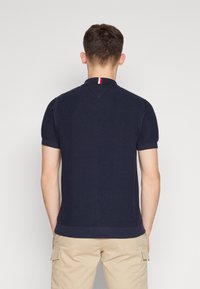 Marineblauw poloshirt met korte mouwen, een gestructureerd patroon en een geribde kraag met rood-witte strepen. Gecombineerd met beige cargoshorts.