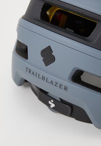 Casco grigio con dettagli neri e lati in rete. Presenta il testo "TRAILBLAZER" e un logo prominente sul fronte. Meccanismo di regolazione a manopola.