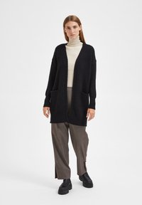 Cardigan noir à devant ouvert, avec deux poches avant et des manches longues, porté par-dessus un col roulé crème et un pantalon marron à rayures.