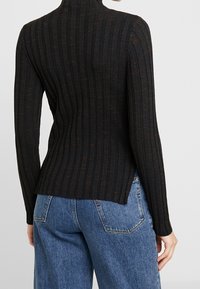 Pull à col roulé côtelé noir en tissu texturé, avec manches longues et fentes latérales, porté avec un jean en denim bleu.