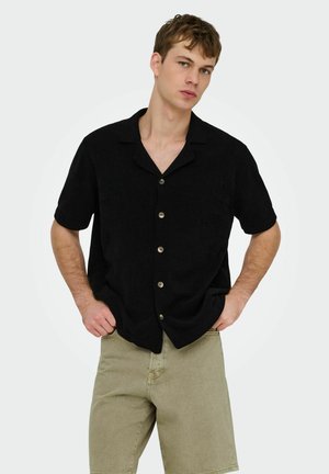 Jeune homme portant une chemise noire à manches courtes boutonnée et un short beige, debout les mains dans les poches sur un fond clair uni.