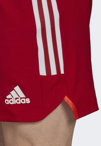 Rode Adidas sportshorts met een wit driebandsontwerp en logo aan de zijkant, gedragen op een harige been tegen een effen achtergrond.
