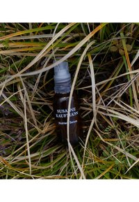 Brązowa szklana butelka z przezroczystą pompą, oznakowana "Serum Odżywcze Susanne Kaufmann", otoczona trawą i suszoną roślinnością.