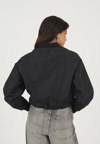 Czarna kurtka bomberka z prążkowanym kołnierzem, prezentująca skróconą sylwetkę i gładką, matową fakturę. W połączeniu z szarymi dżinsami o wysokim stanie.