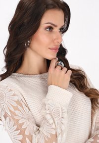 Strickpullover in hellem Creme mit strukturiertem Körper und Ärmel aus floraler Spitze. Das Model trägt einen glänzenden silbernen Ring, der eine sanfte, neutrale Farbpalette präsentiert.