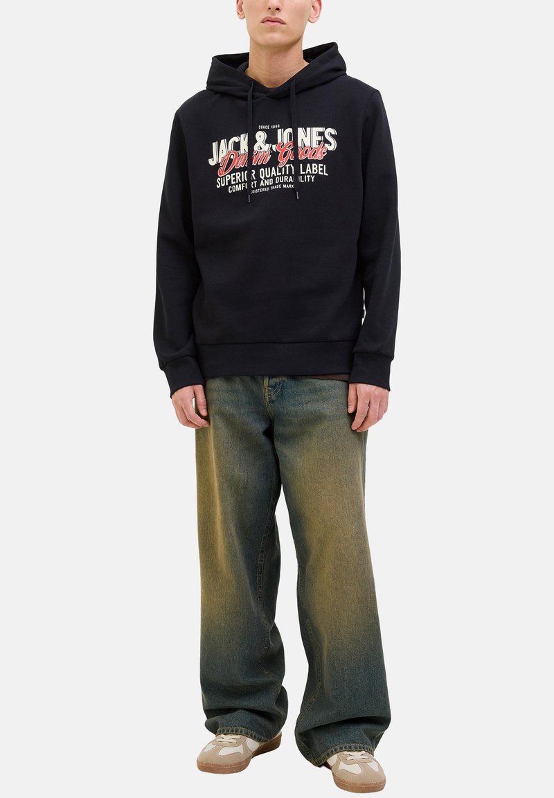 Mann in schwarzem Kapuzenpulli mit weißem und rotem Text, lockeren ausgewaschenen blauen Jeans und beigen und weißen Sneakers, der vor weißem Hintergrund steht.