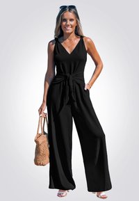 Mono negro sin mangas con detalle de lazada en la cintura, diseño de pierna ancha, fabricado en tejido texturizado. La modelo lleva un bolso de mimbre.