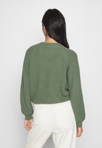 Pull vert en maille avec texture côtelée, manches ballon et col rond, associé à un pantalon blanc taille haute.