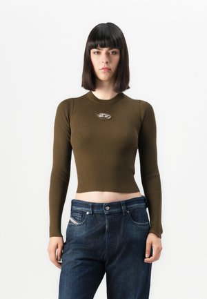 Kvinne med rett, mørkt hår som har på seg en olivengrønn ribbestrikka langermet cropped topp med et lite utsnitt på brystet og mørkeblå jeans mot en hvit bakgrunn.