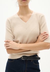 Beige ribbad stickad tröja med V-ringning och korta ärmar, i kombination med mörkblå jeans och ett svart bälte med en silverspänne.