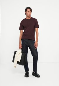 Bordeaux katoenen t-shirt met korte mouwen, gecombineerd met donkere spijkerbroeken. Zwart tote bag met een crèmekleurige trui gevouwen over de handgreep. Zwarte schoenen.
