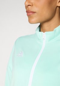 Mint green zip-up jacket made of smooth fabric, featuring a high collar and white zipper. Adidas logo is embroidered in white on the left chest.