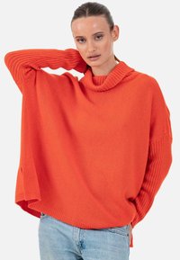 Pull en maille oversize orange vif avec un col bénitier, des manches côtelées, des fentes sur les côtés et une coupe décontractée, associé à un jean bleu clair.