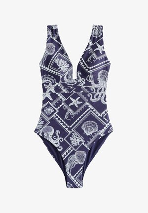 Nő, aki tengerészkék fürdőruhát visel fehér tengeri élőlények és kagylók mintáival, tiszta ég alatt, víz közelében áll.