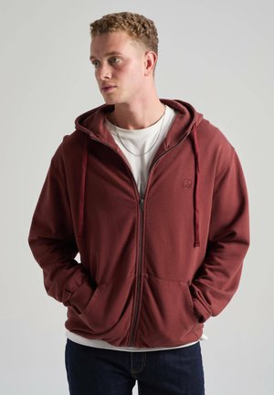 Junger Mann mit lockigem Haar, trägt einen bordeauxroten Kapuzenpullover mit Reißverschluss und ein weißes T-Shirt, Hände in den Taschen, schaut seitlich vor einem einfarbigen Hintergrund.