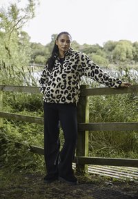 Sudadera de forro polar con estampado de leopardo y cuello con media cremallera, combinada con pantalones de pierna ancha negros. Entorno al aire libre con vegetación de fondo.