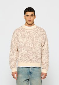 Svetlo béžový sveter so skrúteným ružovým vzorom, s rebrovanými manžetami a lemom a okrúhlym výstrihom, v kombinácii so svetlo modrými džínami.