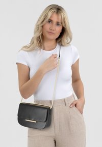 Borsa a tracolla in pelle nera con tracolla a catena, forma rettangolare, dettaglio in metallo dorato sul davanti, texture liscia e design minimalista.