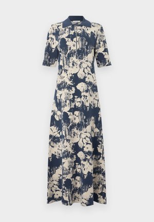 Moteris dėvi tamsiai mėlyną ir kremine abstrakčiu raštu margintą maxi suknelę su apykakle, pusiniais rankovėmis, priekyje užtrauktuku ir juodais plokščiais batais, stovinti viena ranka ant klubo.