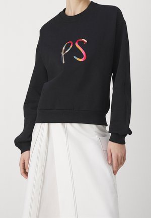 Felpa nera con lettere "ps" colorate sul davanti, indossata con una gonna bianca a vita alta con dettagli a cuciture verticali e tasca laterale.