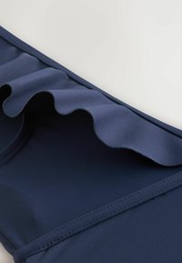 Tengerészkék ruhadarab sarka, egy sima fodor részlettel az él mentén, fehér háttéren.