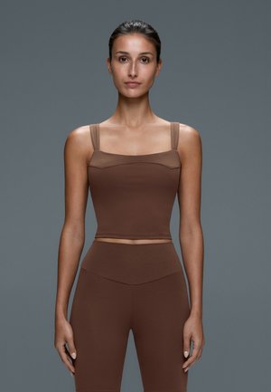 Giovane donna con capelli scuri legati indietro indossa un crop top marrone aderente e leggings a vita alta abbinati su uno sfondo grigio.