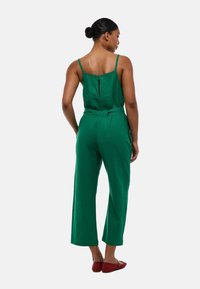 Combinaison en lin vert avec un corsage ajusté, des bretelles spaghetti réglables, une fermeture éclair dans le dos et un pantalon évasé. Associée à des chaussures rouges.