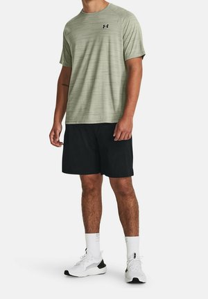 Mies pukeutuneena vaaleanvihreään lyhythihaiseen urheilupaitaan, mustiin shortseihin, valkoisiin sukkiin ja valkoisiin lenkkareihin, seisoo yksiväristä taustaa vasten.