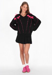 Schwarzer übergroßer Pullover mit rosa Schleifenakzenten und kontrastierenden Nähten, kombiniert mit einem schwarzen plissierten Rock und rosa Turnschuhen.